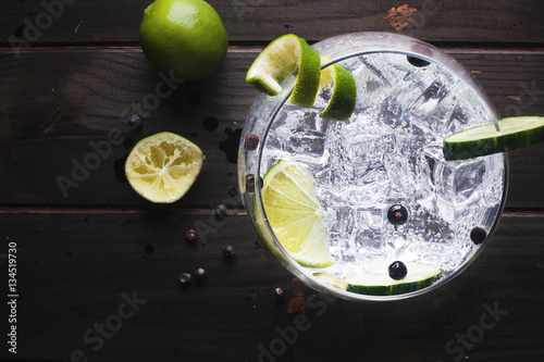 Glass of gin tonic with ice, cucumber, lime over a dark wood table photo
