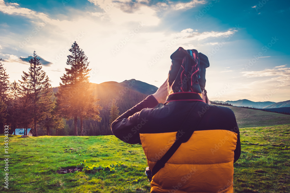 Fototapeta premium Nature photographer tourist with camera taking a photo in the mountains. Dreamy sunset landscape, spring green meadow and mountain top in the bsckground. Back view. Beautiful nature landscape. Travel