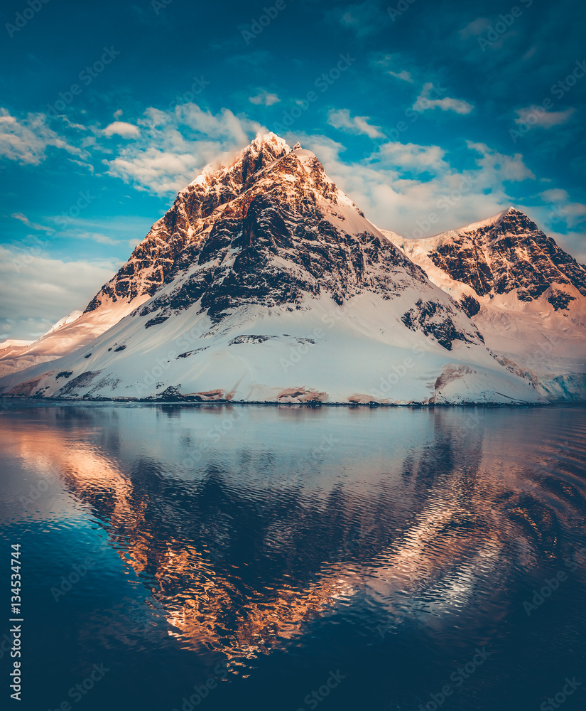 Antarctic landscape with snow covered mountains reflected in ocean water. Sunset warm light on the mountain peak, blue cloudy sky in the background. Beautiful nature landscape. Travel background.