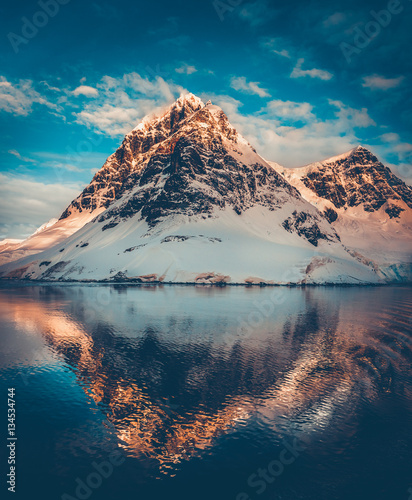 Antarctic landscape with snow covered mountains reflected in ocean water. Sunset warm light on the mountain peak, blue cloudy sky in the background. Beautiful nature landscape. Travel background. © Goinyk