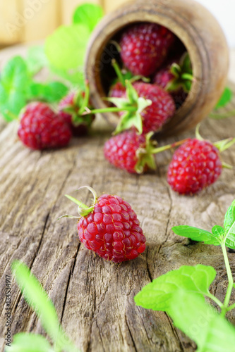 Red fresh raspberries on rough rustic wooden background. Cup with natural ripe organic berries with peduncles and raw green mint on table. Vertical  close up.