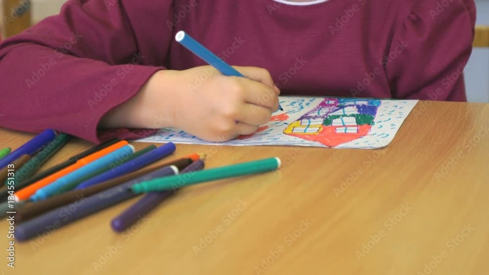 Kid sitting at the desk draws the image on the paper sheet using the colored felt pens. Close-up Kid sitting at the desk draws the image on the paper sheet using the colored felt pens. Close-up