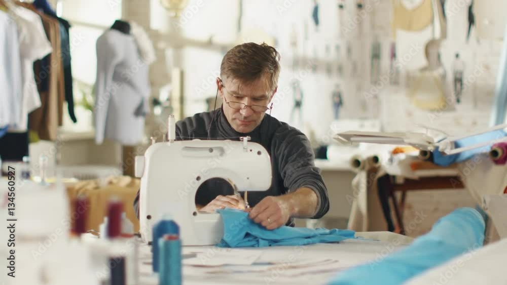 Male Fashion Designer/ Dressmaker Working on a Sewing Machine in Him Sunny Studio. Various Sewing items and Fabrics Laying Around Him, Visible Sketches Pinned to the Wall. Shot on RED EPIC 4K (UHD). Male Fashion Designer/ Dressmaker Working on a Sewing Machine in Him Sunny Studio. Various Sewing items and Fabrics Laying Around Him, Visible Sketches Pinned to the Wall. Shot on RED EPIC 4K (UHD).