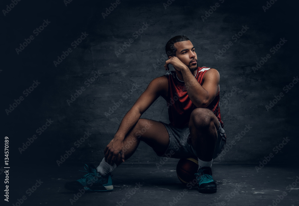 A black basketball player sits on a floor and holds basket ball. A black basketball player sits on a floor and holds basket ball.