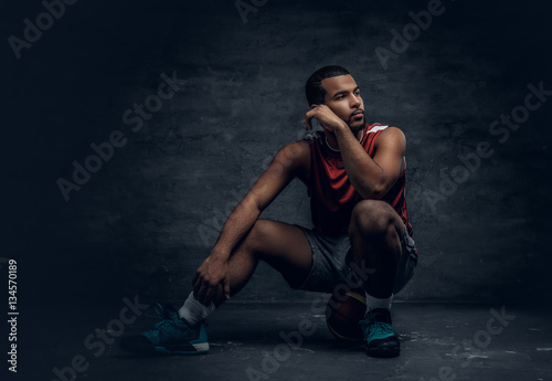 A black basketball player sits on a floor and holds basket ball.