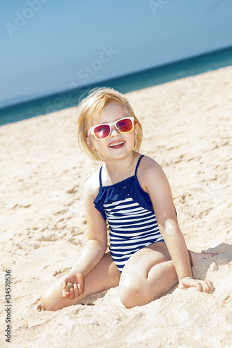 Happy girl in striped swimsuit playing with sand on white beach
