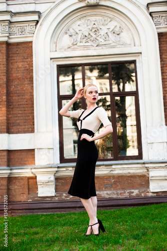 Happy Fashion blonde european elegant Woman with red lips and white skin Standing at the Old Red Brick building on green grass. Beautiful Female in formal black and white dress wearing sunglasses. 