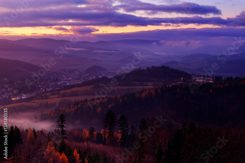 Small town Jesenik with spa resort in the mountain forest. Czech early morning landscape with beautiful sunrise with pink and violet clouds. Autumn trees with fog, Jeseniky mountains. End of night.