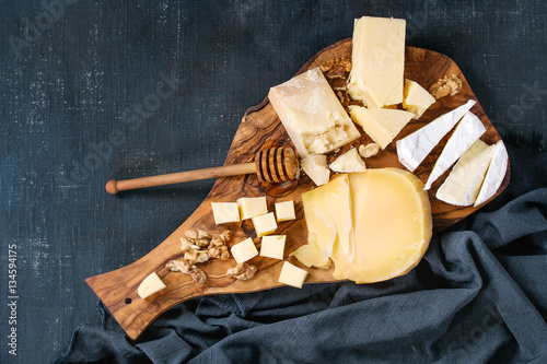 Cheese plate. Assortment of cheese with walnuts, honey from honey dipper on olive wood serving board with textile over dark blue canvas as background. Top view with space.
