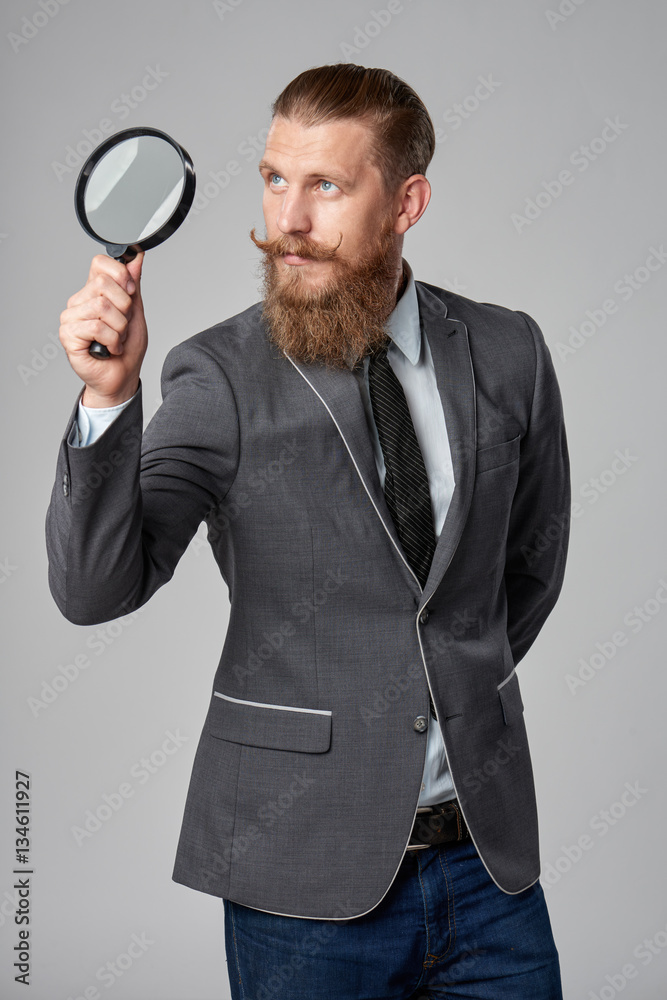 Serious hipster business man with beard and mustashes in suit looking up through magnifying glass, over grey background Serious hipster business man with beard and mustashes in suit looking up through magnifying glass, over grey background