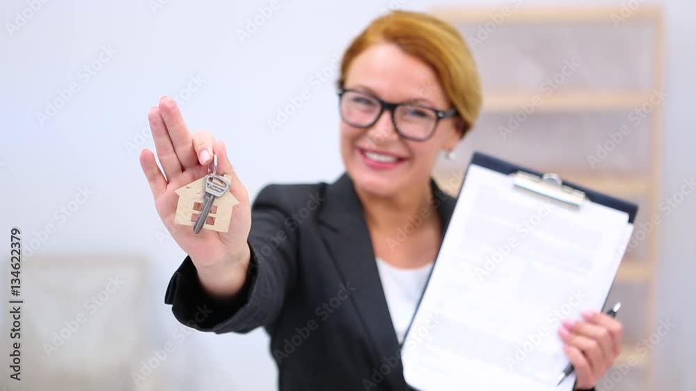 Happy smiling realtor lady in glasses offering new key for new apartment or flat in newly built house located in beautiful residential area. Happy smiling realtor lady in glasses offering new key for new apartment or flat in newly built house located in beautiful residential area.