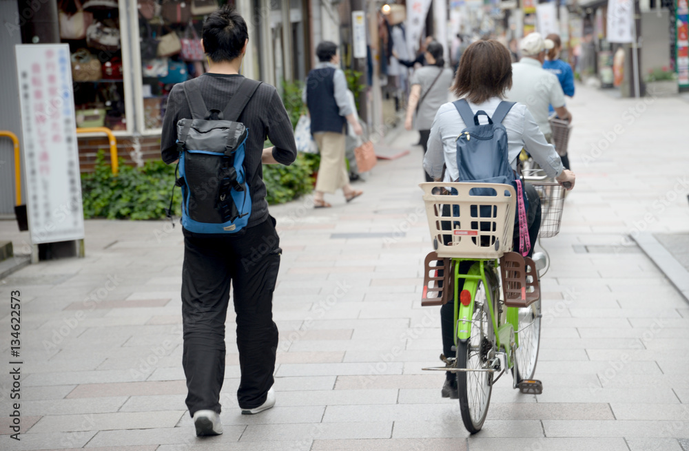 Idący pieszo młody mężczyzna z plecakiem oraz kobieta z plecakiem jadąca na rowerze z zamontowanym koszykiem do przewożenia dziecka - obrazy, fototapety, plakaty 