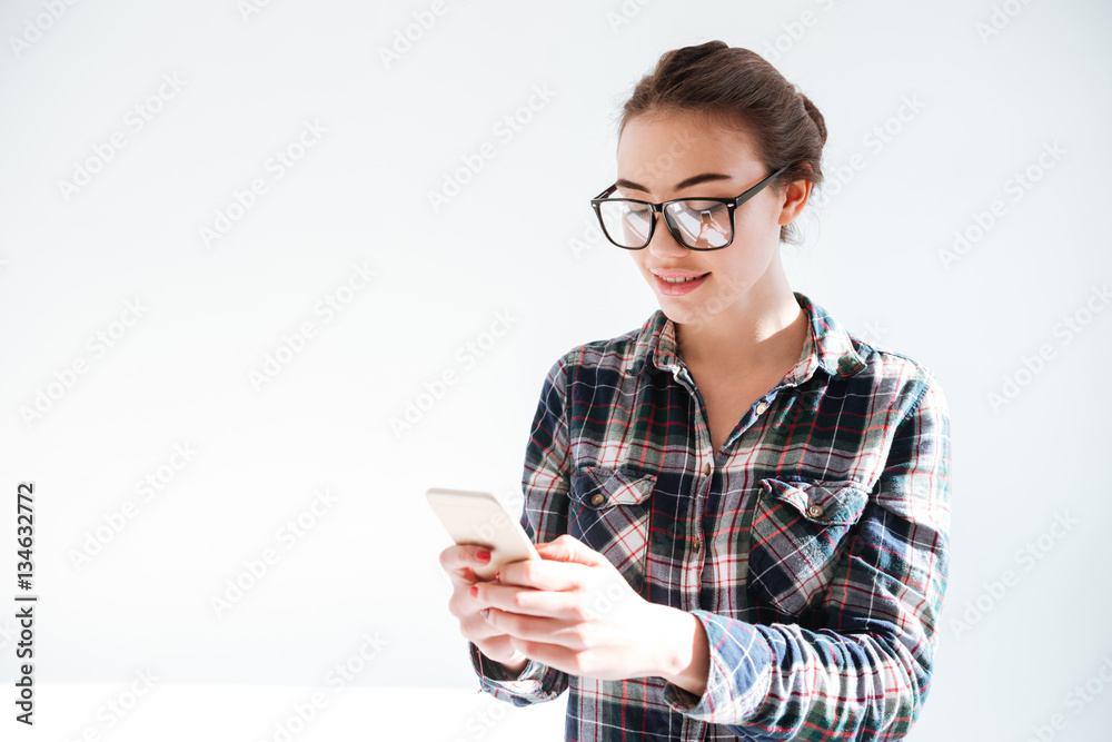 Smiling young woman in glasses standing and using mobile phone