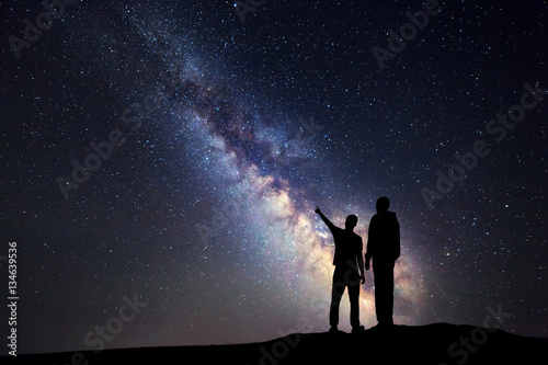 Silhouette of a father and a son who pointing finger in night starry sky on the background of Milky Way. Family. Colorful night landscape. Beautiful Universe. Space. Travel background