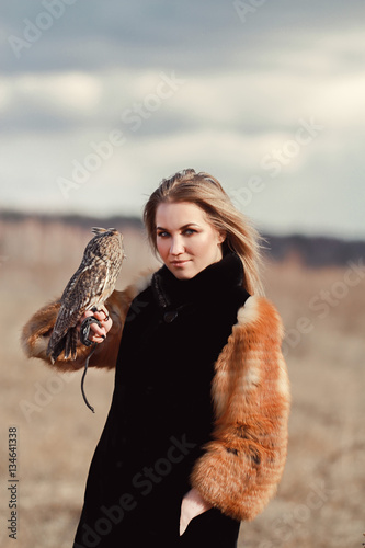 Beautiful woman in a fur coat with an owl on his arm. Blonde with long hair in nature holding a owl. Romantic delicate image of a girl