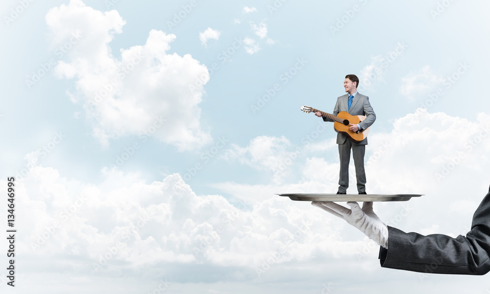 Businessman on metal tray playing acoustic guitar against blue sky background Businessman on metal tray playing acoustic guitar against blue sky background