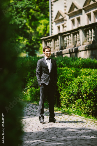 Groom in classy black tuxedo stands on path along green bushes