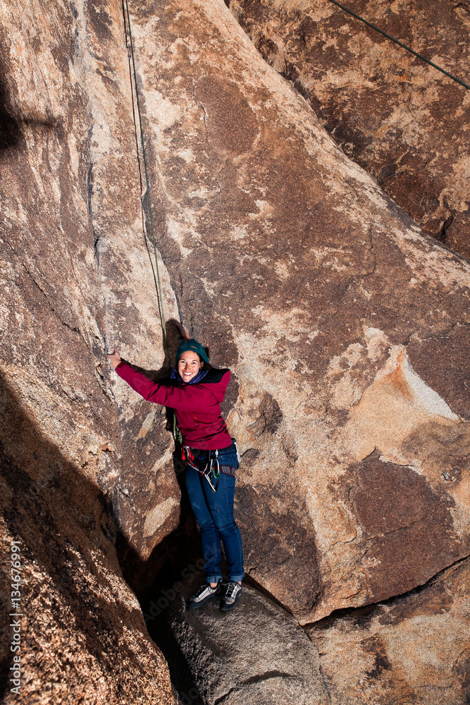 Young caucasian woman dressed for cold weather rock climbing in the desert climbs a cliff and smiles at the viewer