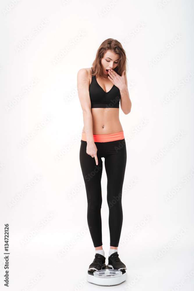Full length of astonished young sportswoman standing on weighting scale Full length of astonished young sportswoman standing on weighting scale