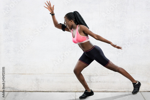 Full length action portrait of muscular african female athlete in running position. Side view of young woman shouting at take off. Girl in sportswear against light grey concrete background. 
