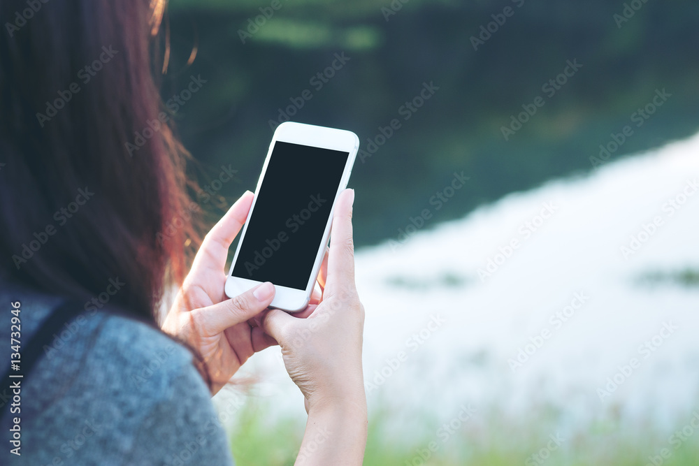 Mockup image of a woman using smart phone with blank black screen at outdoor and lake nature background