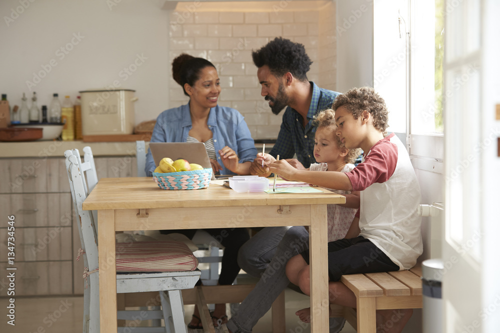 Children Paint At Kitchen Table As Parents Look At Laptop Children Paint At Kitchen Table As Parents Look At Laptop