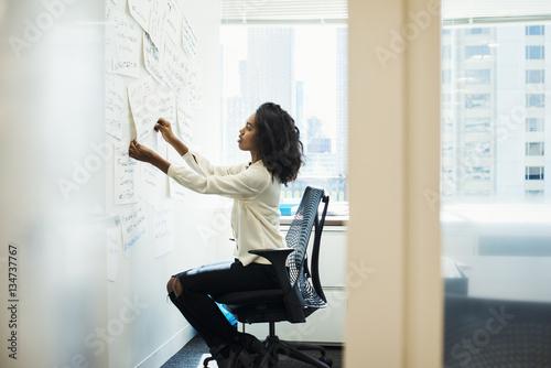 A woman sitting on a chair in an office arranging pieces of paper pinned on a whiteboard. photo