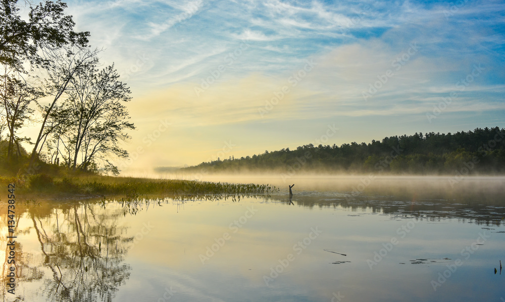 Fototapeta premium Brilliant and bright mid-summer sunrise on a lake. Warm water and cooler air at daybreak creates misty fog patches. Still water along a calm, quiet Ontario lakeside. 