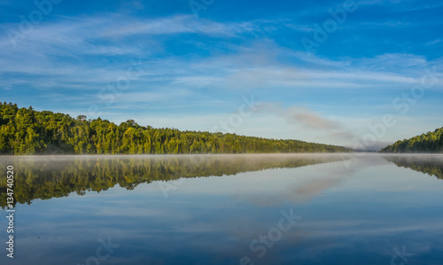 Bright mid-summer blue sky  misty morning in the middle of Corry lake.   Warm water and cooler air at daybreak creates misty patches along shoreline.  Still water along a calm  quiet lakeside.