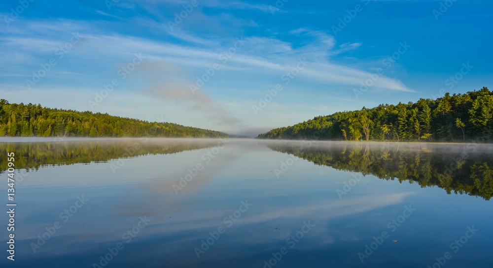 Bright mid-summer blue sky, misty morning in the middle of Corry lake.   Warm water and cooler air at daybreak creates misty patches along shoreline.  Still water along a calm, quiet lakeside.