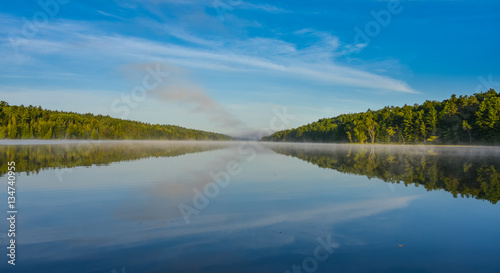 Bright mid-summer blue sky, misty morning in the middle of Corry lake. Warm water and cooler air at daybreak creates misty patches along shoreline. Still water along a calm, quiet lakeside.