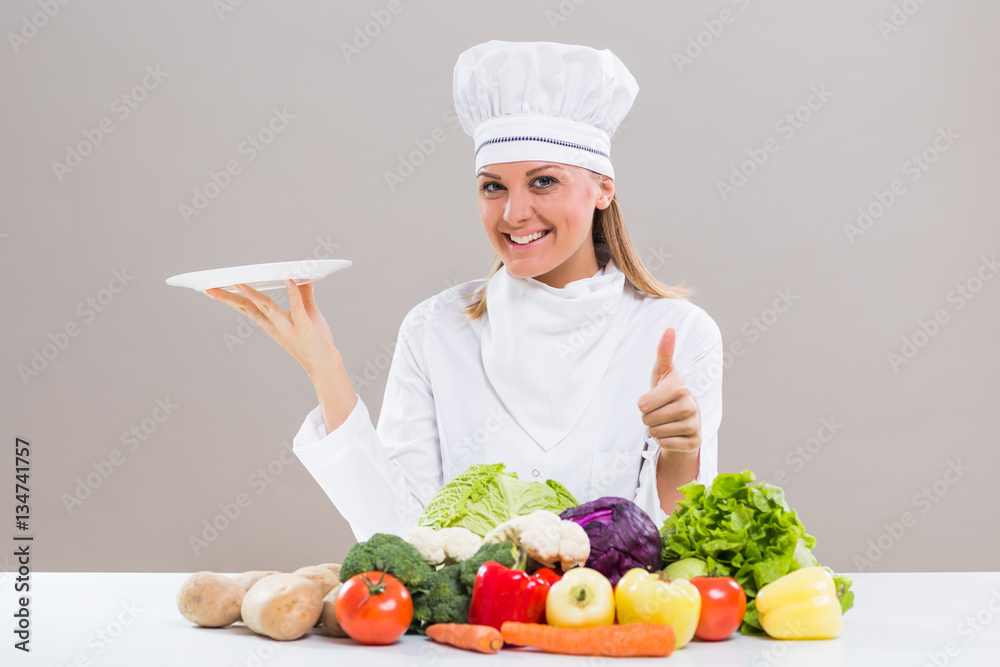 Female chef surrounded with vegetables holding plate showing thumb up Female chef surrounded with vegetables holding plate showing thumb up