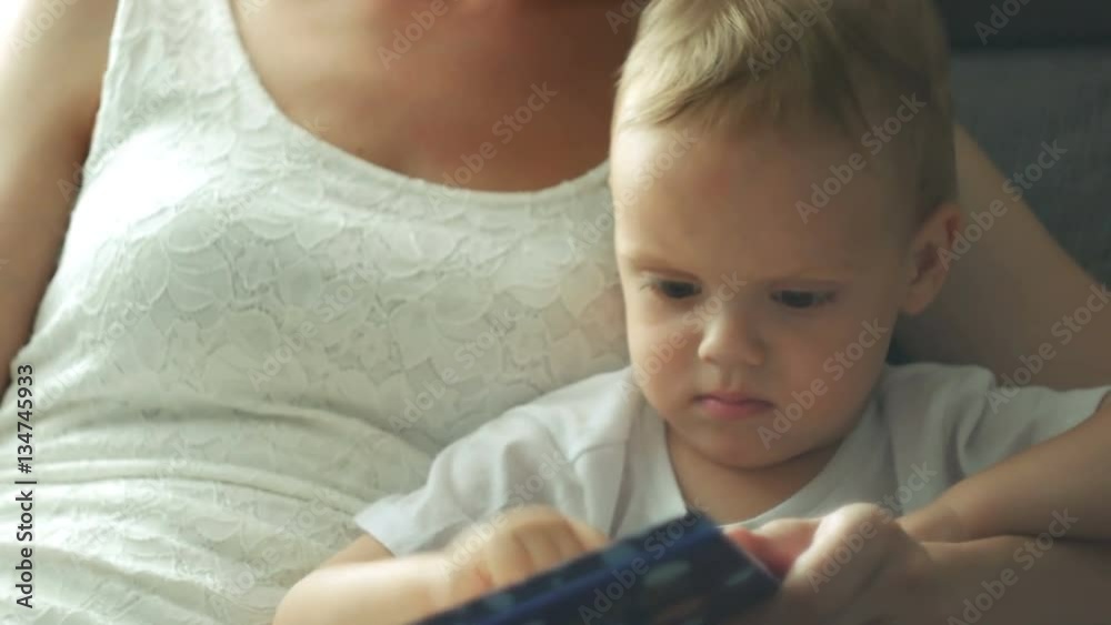 Mother and her two years old baby boy are reading book sitting in armchair in sunny room Mother and her two years old baby boy are reading book sitting in armchair in sunny room