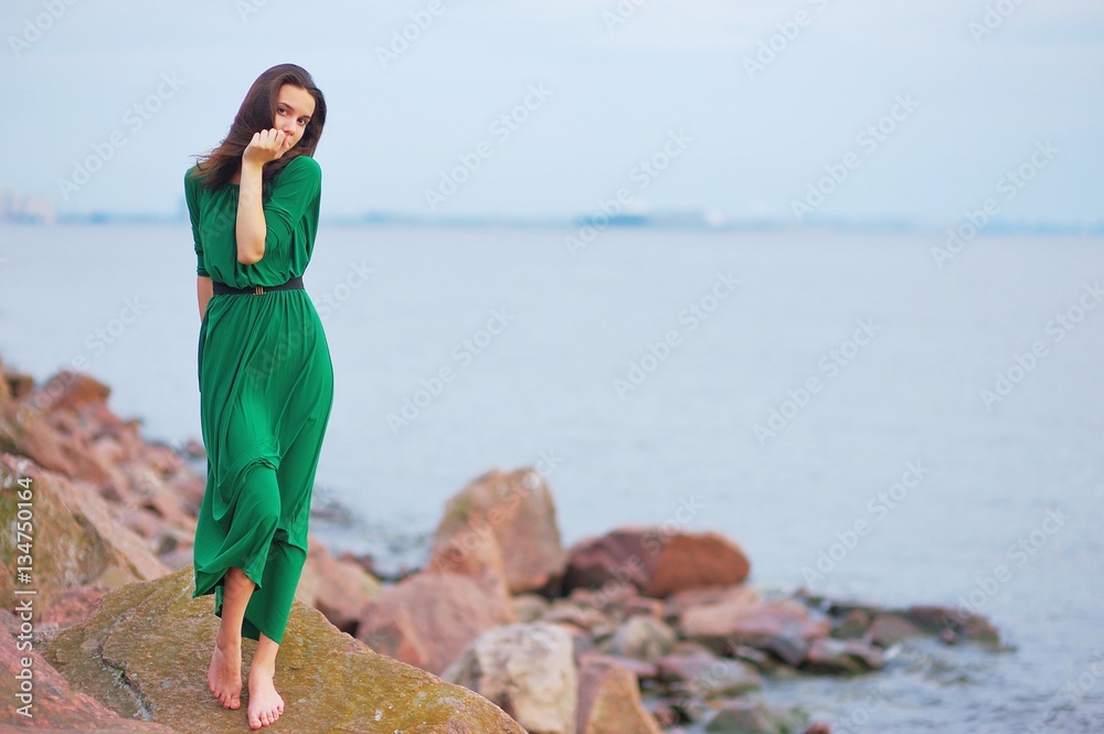 beautiful woman in a green dress looks at the evening sun. she stands barefoot on the rocks by the sea.