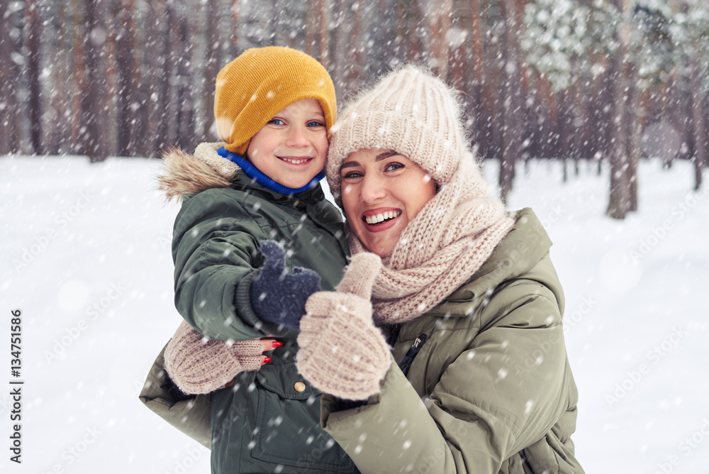 Smiling mom hugging her son under the snowfall in the forest