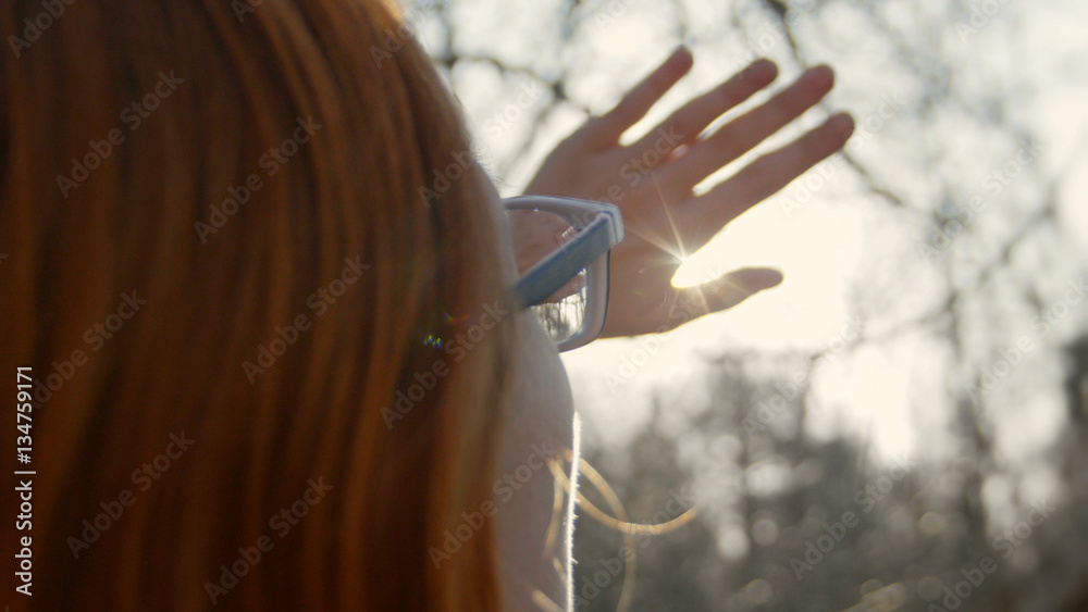 beautiful girl with red hair wearing glasses in the spring park looks at the sun