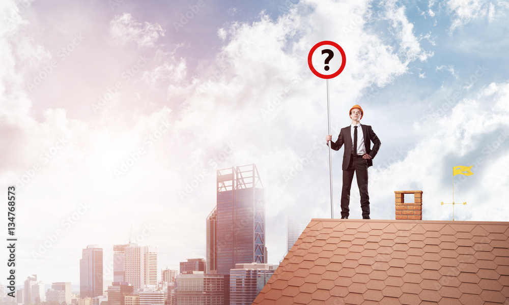 Businessman on brick house roof showing banner with question mark. Mixed media Businessman on brick house roof showing banner with question mark. Mixed media
