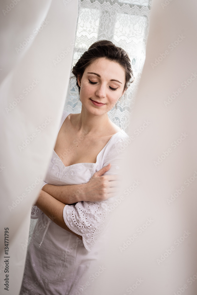 Beautiful woman standing by the window between curtains on natural light background Beautiful woman standing by the window between curtains on natural light background