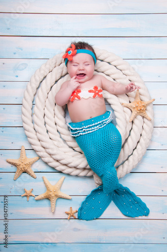 The newborn girl is crying in a mermaid suit the ropes on wooden boards.