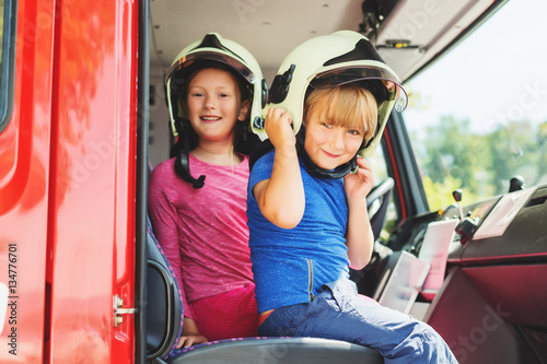 Two cute kids playing in fire truck, pretending to be firefighters, open doors day at fire station. Future profession for children. Educational program for schoolkids