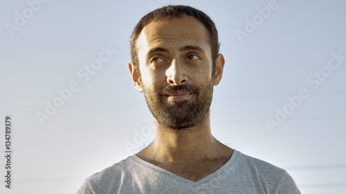 Tanned man with beard and in gray t-shirt smiles with teeth. Cheerful person. Human under sunlight. Joy, happiness and consent concept. Close-up. Front view.