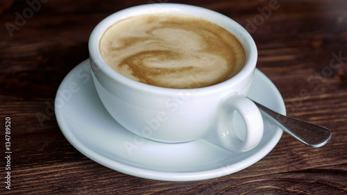 White cup with coffee, saucer and teaspoon on wooden table surface. Close-up.