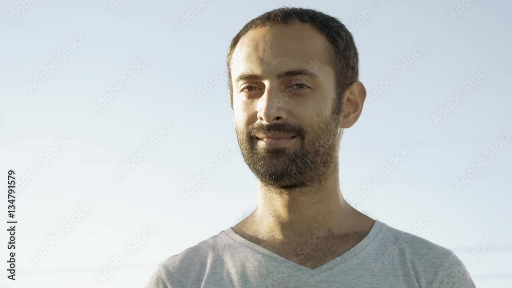 Tanned man with beard and in gray t-shirt smiles with teeth and nods head. Cheerful person. Human under sunlight. Joy, happiness and consent concept. Close-up. Front view. Tanned man with beard and in gray t-shirt smiles with teeth and nods head. Cheerful person. Human under sunlight. Joy, happiness and consent concept. Close-up. Front view.