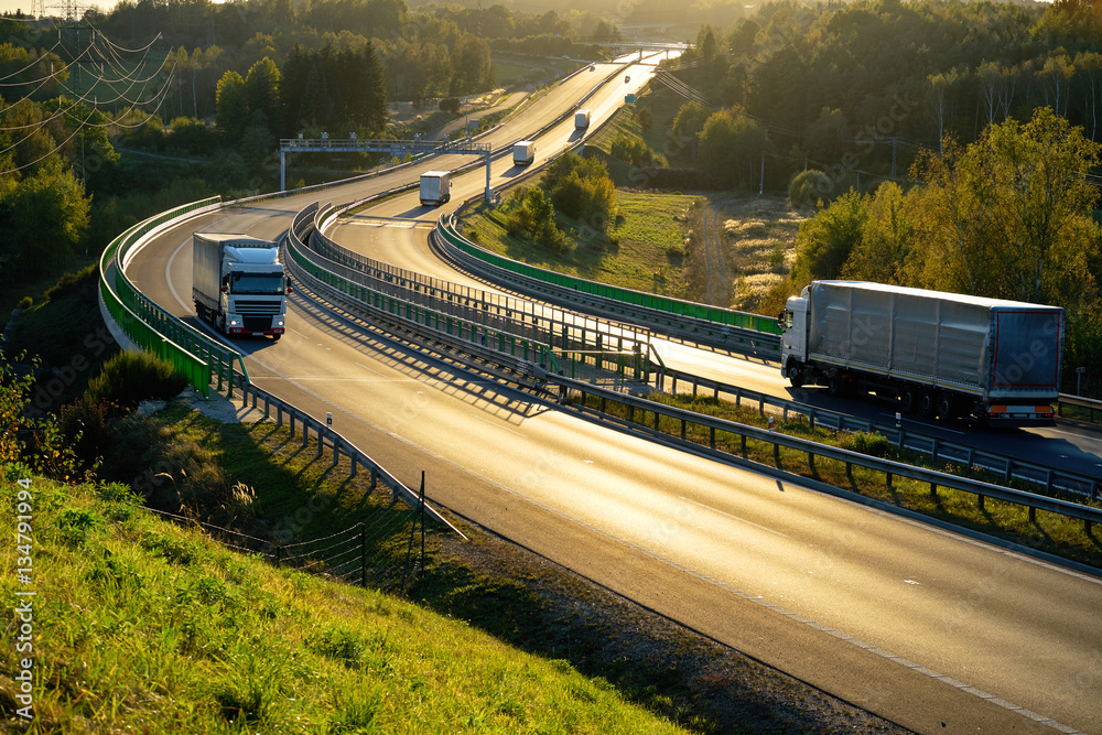 White trucks passing through tollgates on the highway winding through the wooded valley in backlit. View from above.