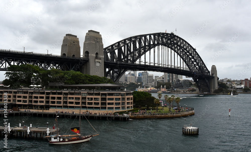Sydney, Australia - January 26, 2017. Tall ship with aboriginal flag sailing out from Circular Quay on Australia Day. Harbour Bridge with cloudy sky in the background.