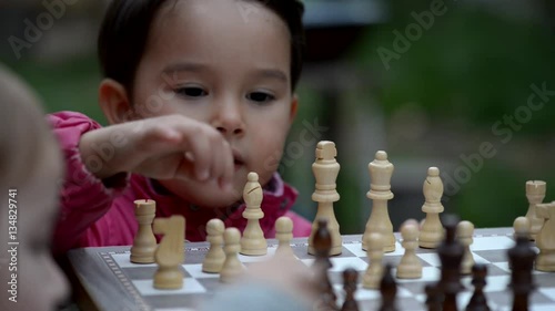 Little child plays wooden chess in park, people walk around 