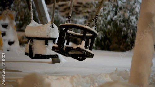 In the frame there is a lonely swing swaying from side to side in abandoned winter park covered with snow illuminated with the light of street lamp. 