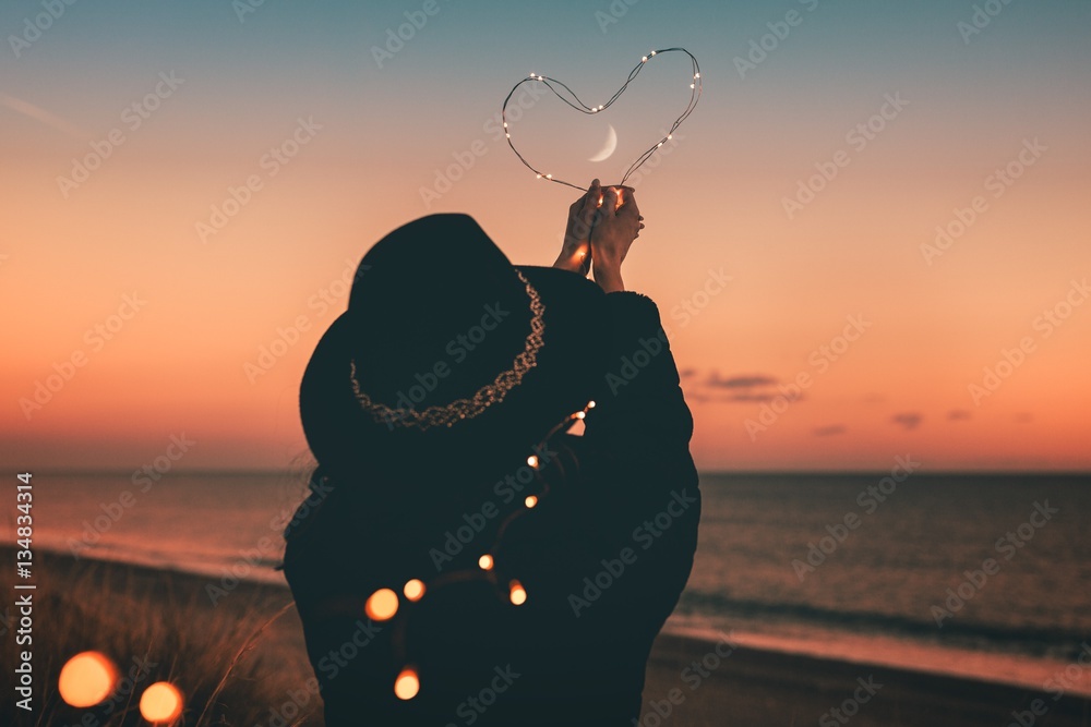 Cuore tenuto in mano da una ragazza vestita di nero con lo sfondo del mare durante il tramonto - obrazy, fototapety, plakaty 