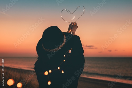 Cuore tenuto in mano da una ragazza vestita di nero con lo sfondo del mare durante il tramonto