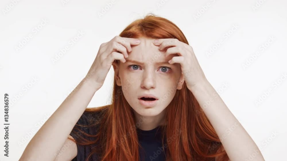 Young beautiful Caucasian red-haired girl peering at camera with displeasure on white background Young beautiful Caucasian red-haired girl peering at camera with displeasure on white background